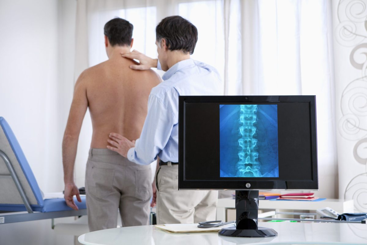  Image of a male patient getting a neck and back examination standing behind a computer screen with an image of his spine.