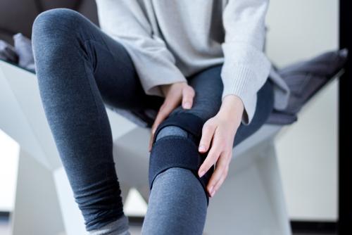 Young woman in gray sweatshirt and leggings holds her braced knee recovering from torn meniscus surgery. 