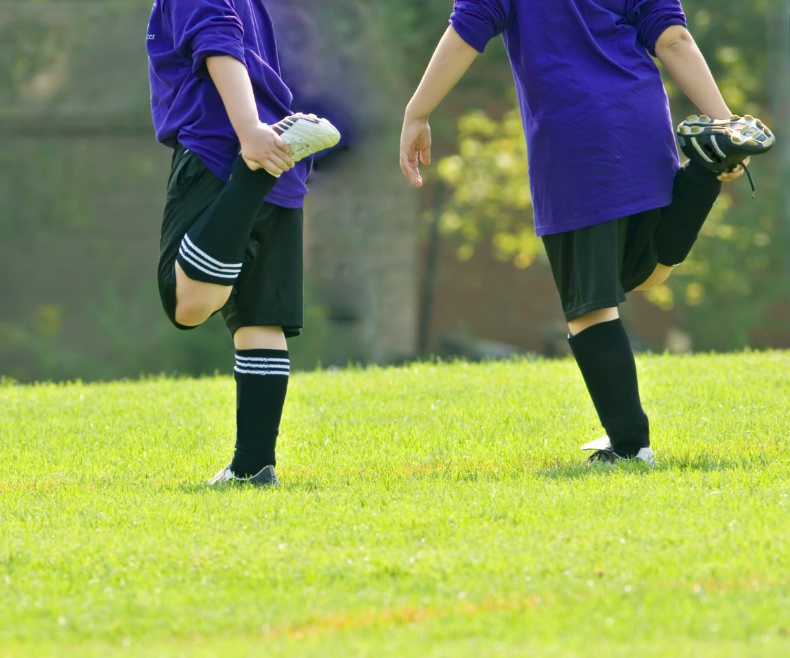  Two soccer players stand on the field and warm up by stretching their legs. 