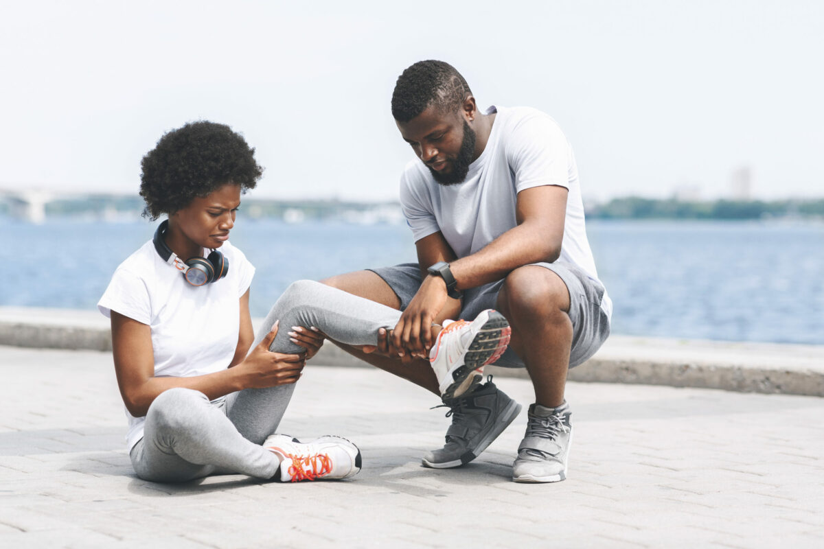 A young woman sits on the ground outside while a young man examines her sprained ankle. 