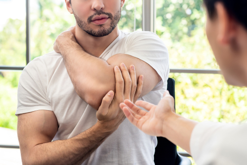 A fit male holds the underside of his elbow during a consultation for rotator cuff tendonitis with a male orthopedist.