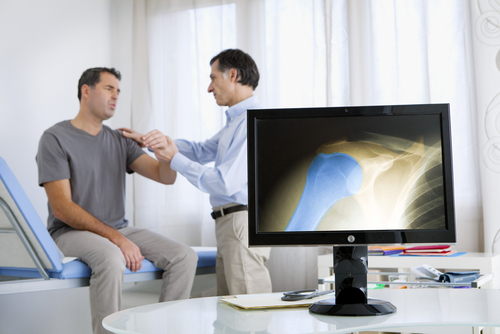 A computer screen highlights an arthritic shoulder, while a doctor examines the stiff, painful shoulder of the male patient.