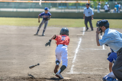 Boy in red baseball uniform drops bat and runs to first base while a boy in a blue baseball uniform guards the base. 