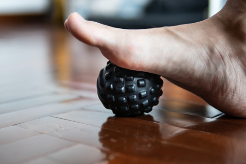 A male’s foot is pictured practicing a foot exercise with a small golf-ball-sized ball between his foot and hardwood floor.