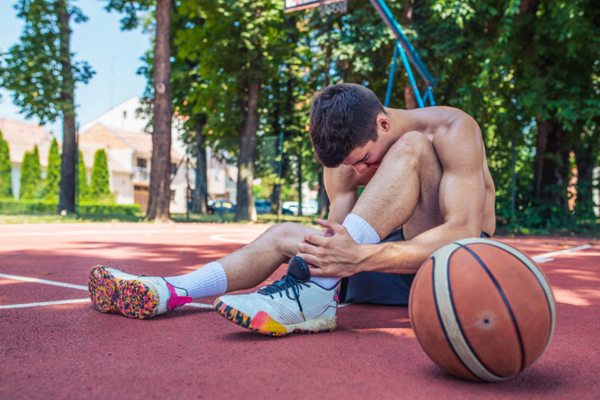  Basketball player on an outdoor court sitting and holding his ankle in pain. 