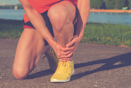 A female athlete wearing yellow athletic shoes squats down to hold her injured ankle during an outdoor run. 