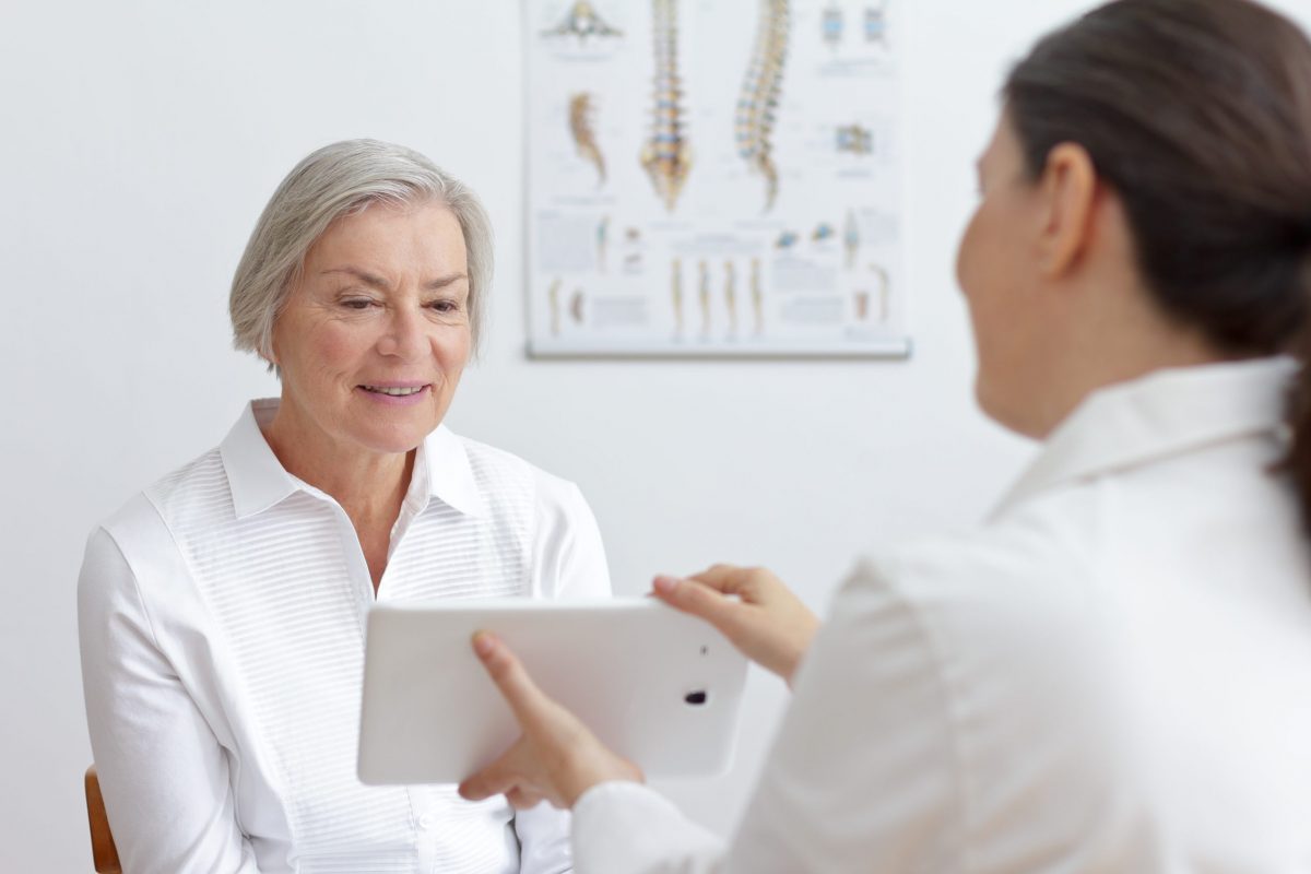 A female orthopedic bone doctor shares an image on her tablet with a female patient, a poster of the spine in background.