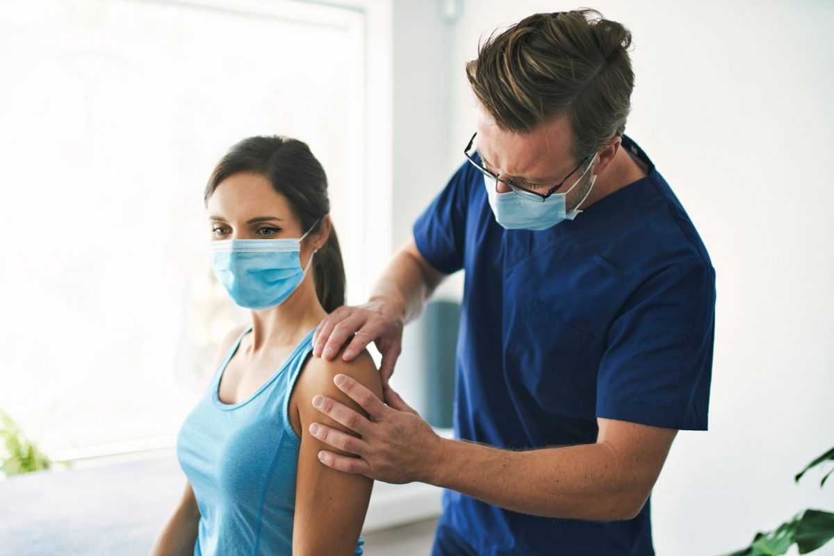 A male orthopedic doctor wearing a mask examines the shoulder of female patient wearing mask, for possible PRP therapy.