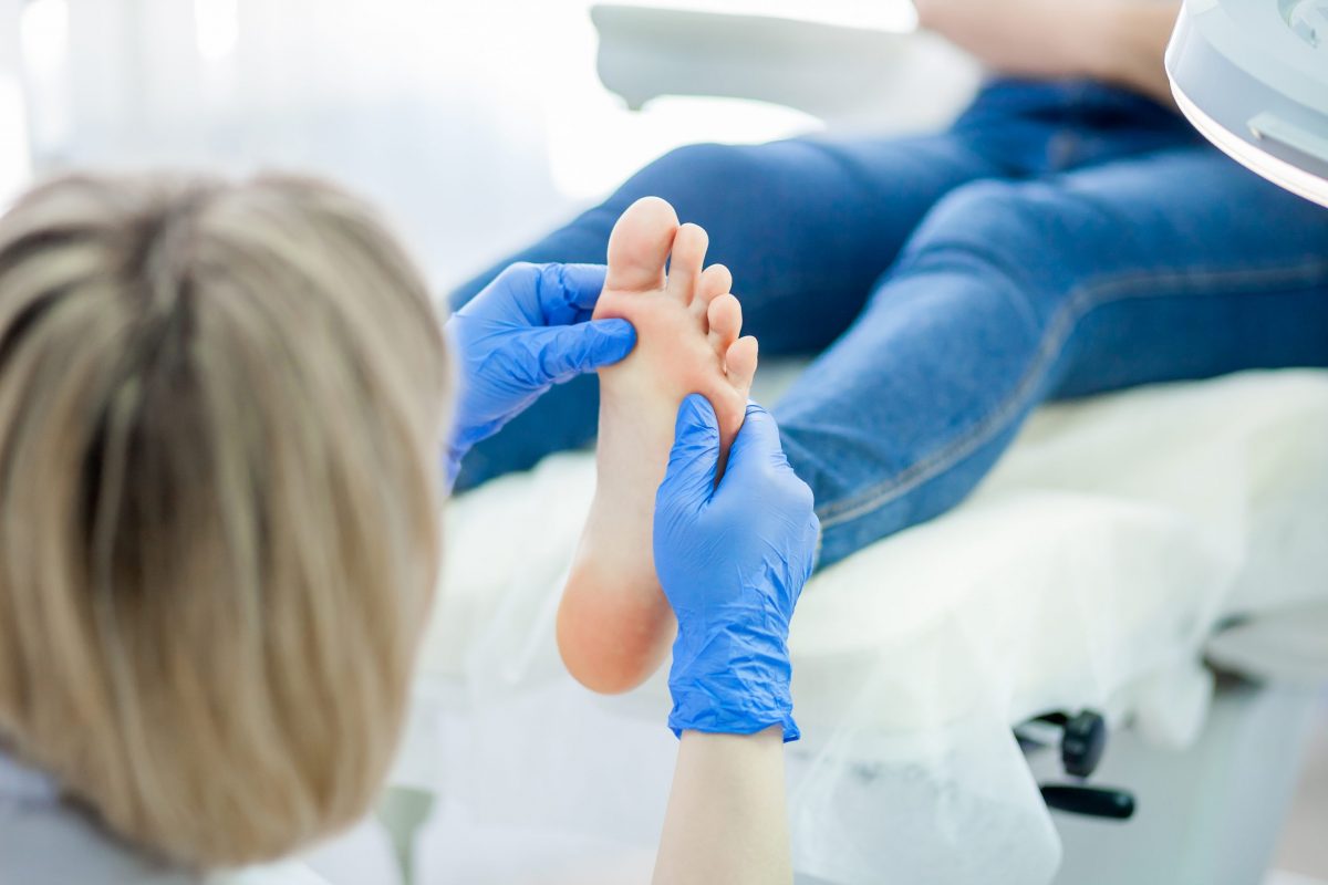 A female podiatrist with blonde hair examines the foot of a young female patient wearing jeans.