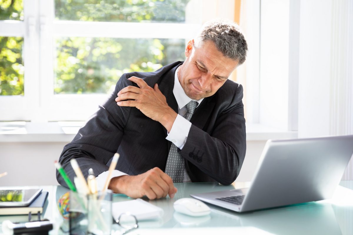 Mature man in gray suit holds his shoulder in pain while working in an office. 