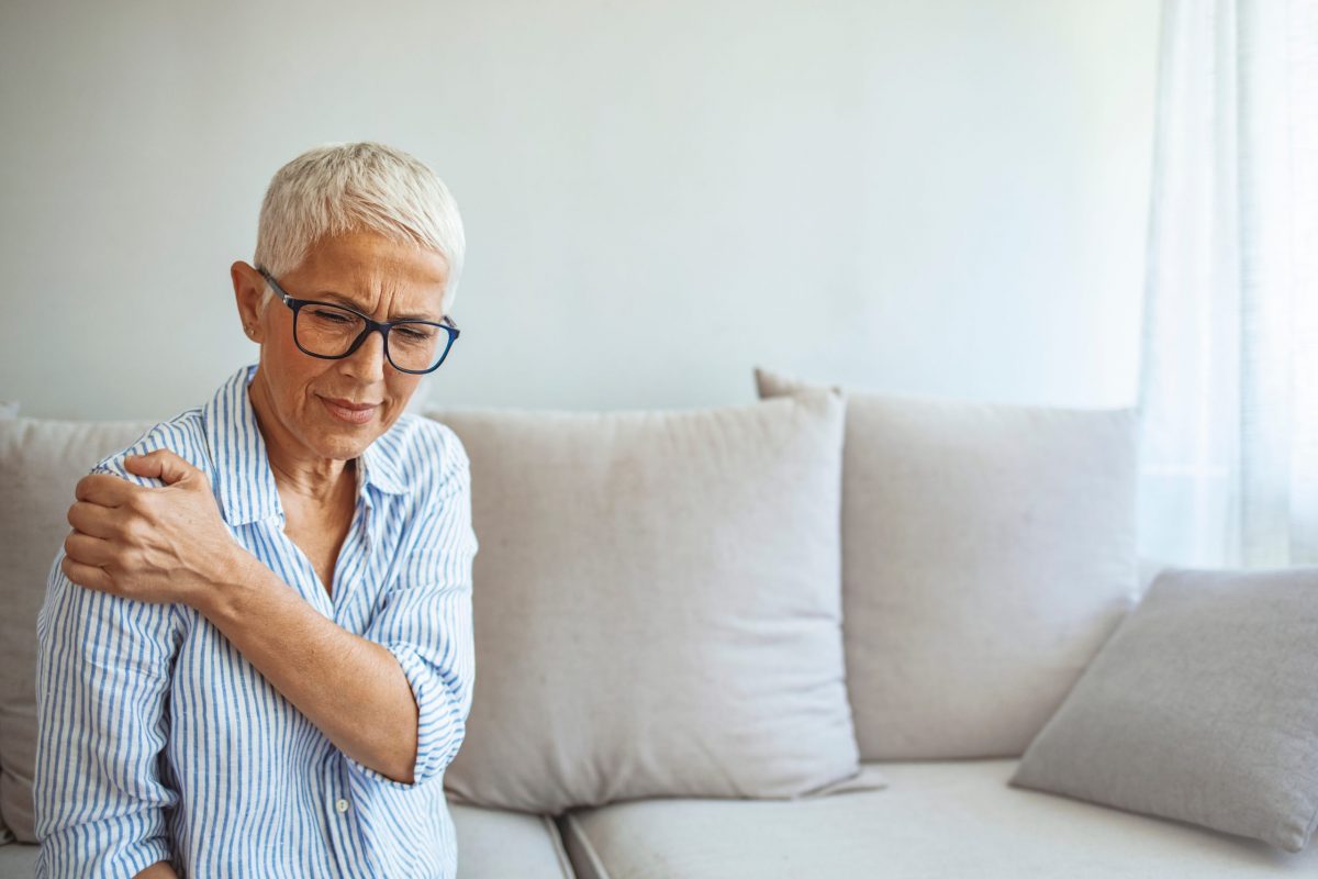 A mature woman sitting on her couch holds her elbow in pain. 