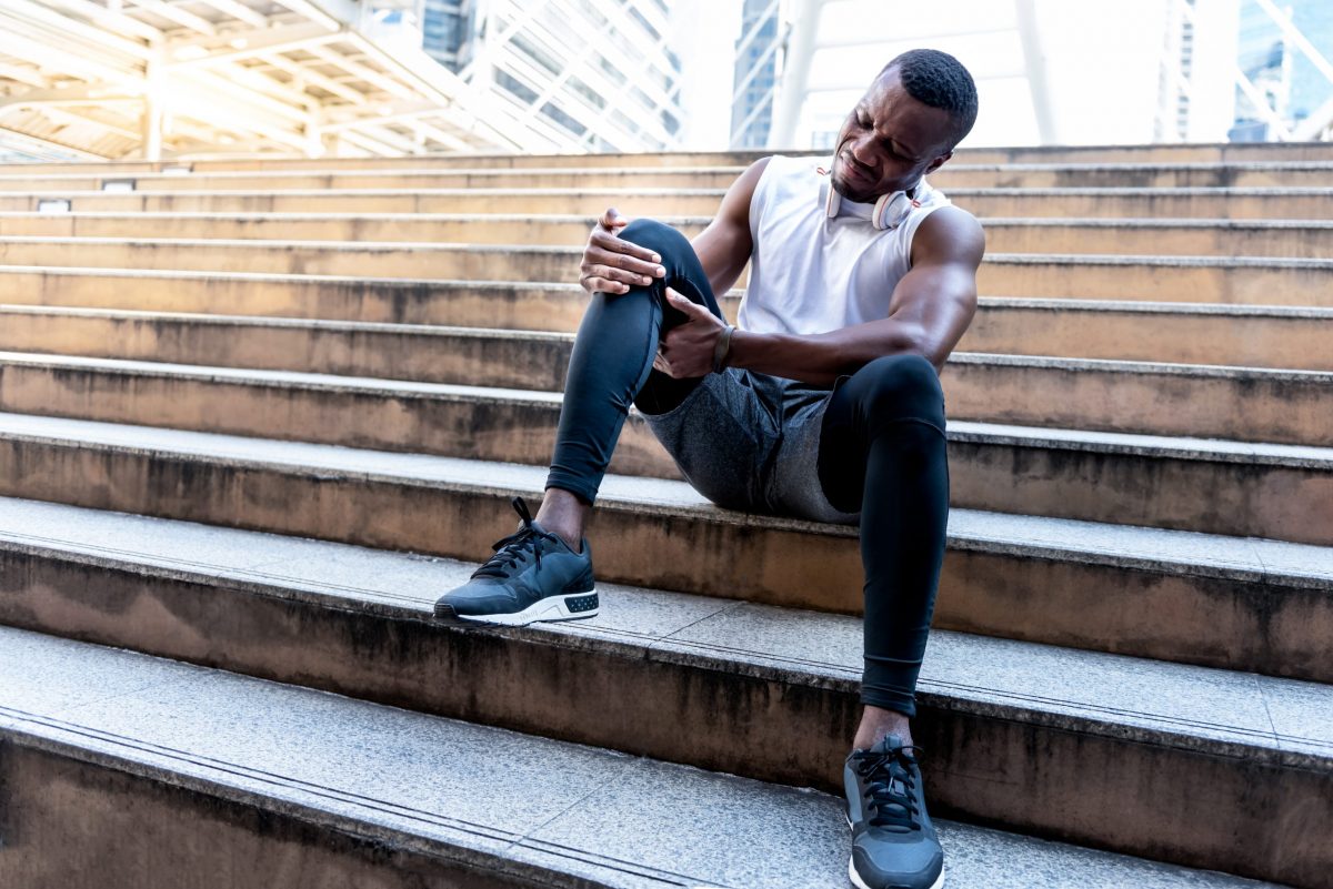 Black male athlete in workout gear sits on concrete steps holding his knee pain. 