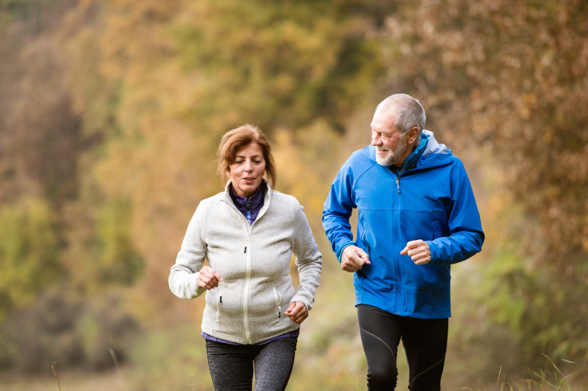 Beautiful senior couple man and woman in jackets going for a jog outside in sunny autumn forest