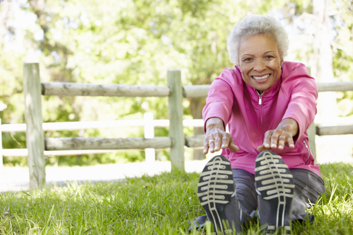 Smiling woman in pink jacket stretches before her workout in a sunny park.