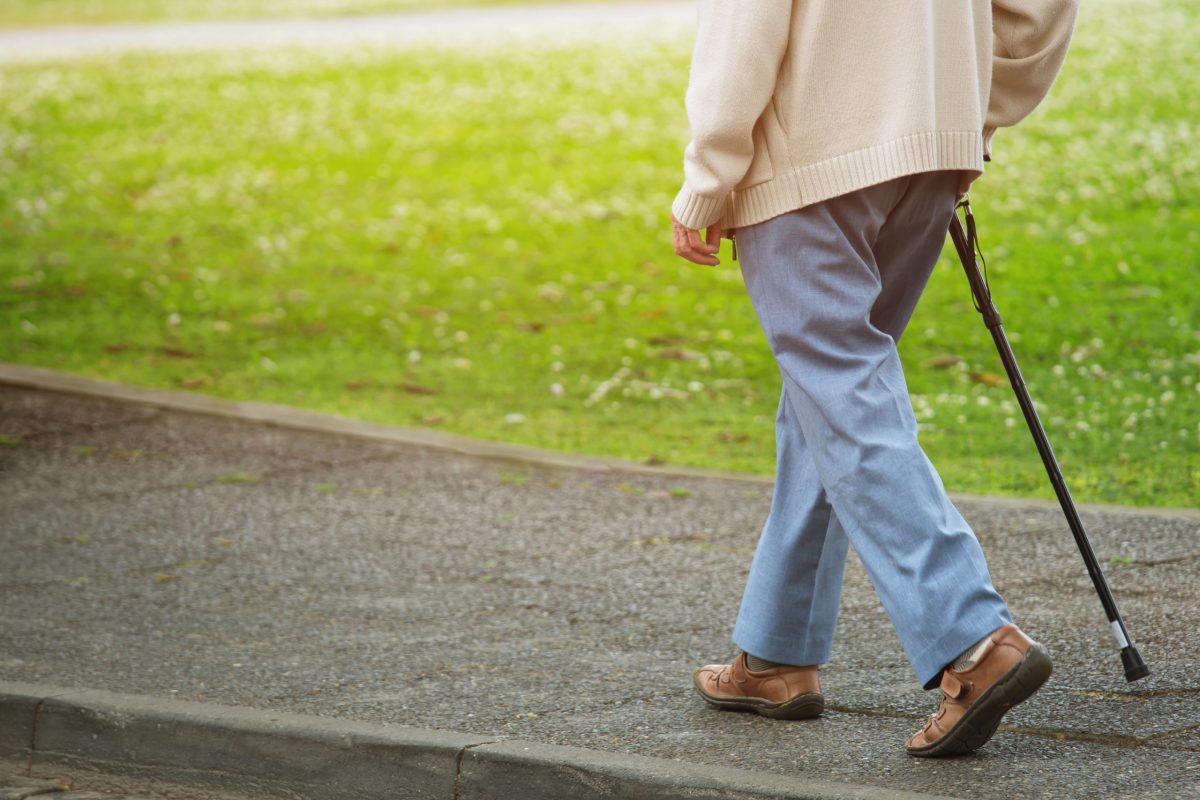  An older adult walks along a sidewalk with a cane. 