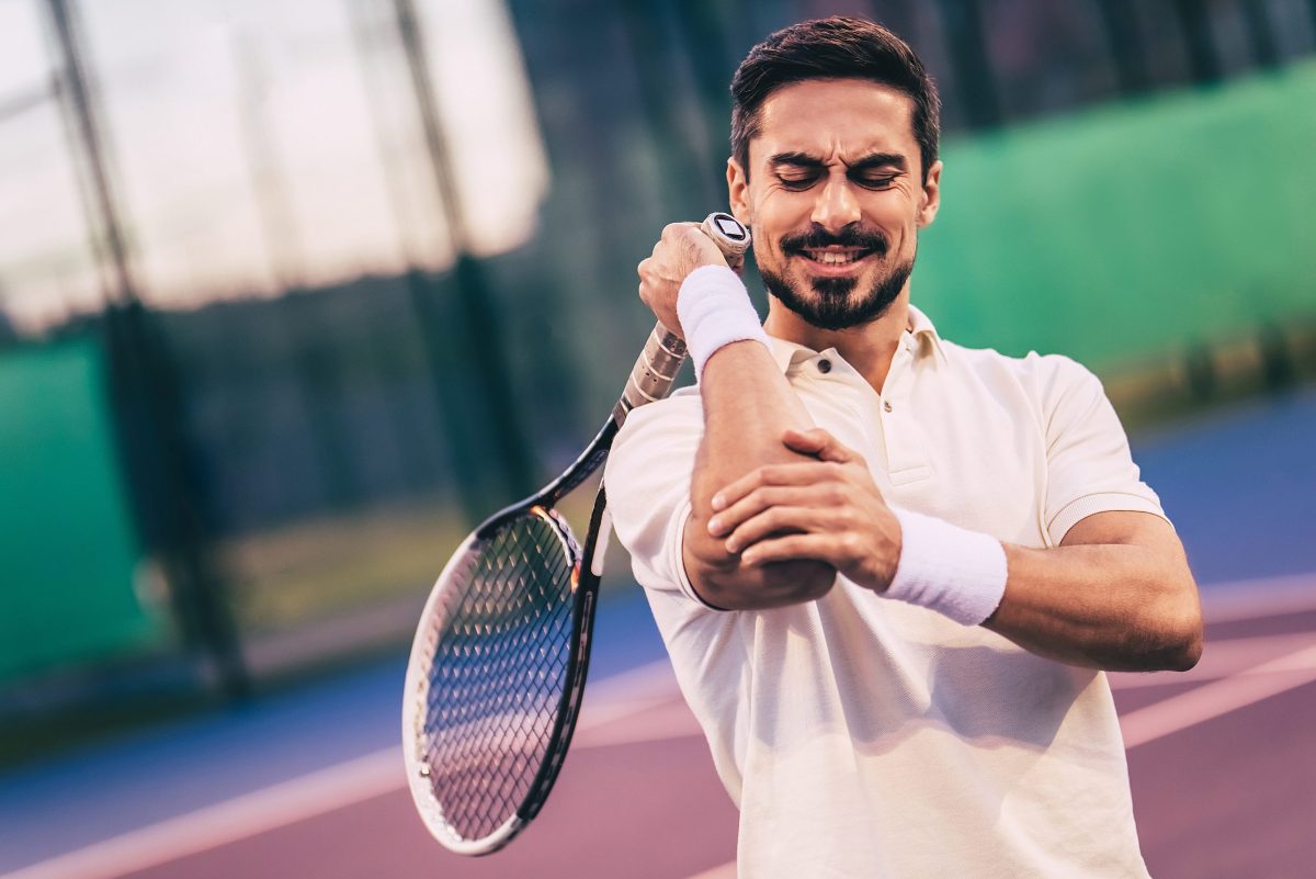 A male tennis player with a tennis racket in hand in white clutches his elbow in pain 