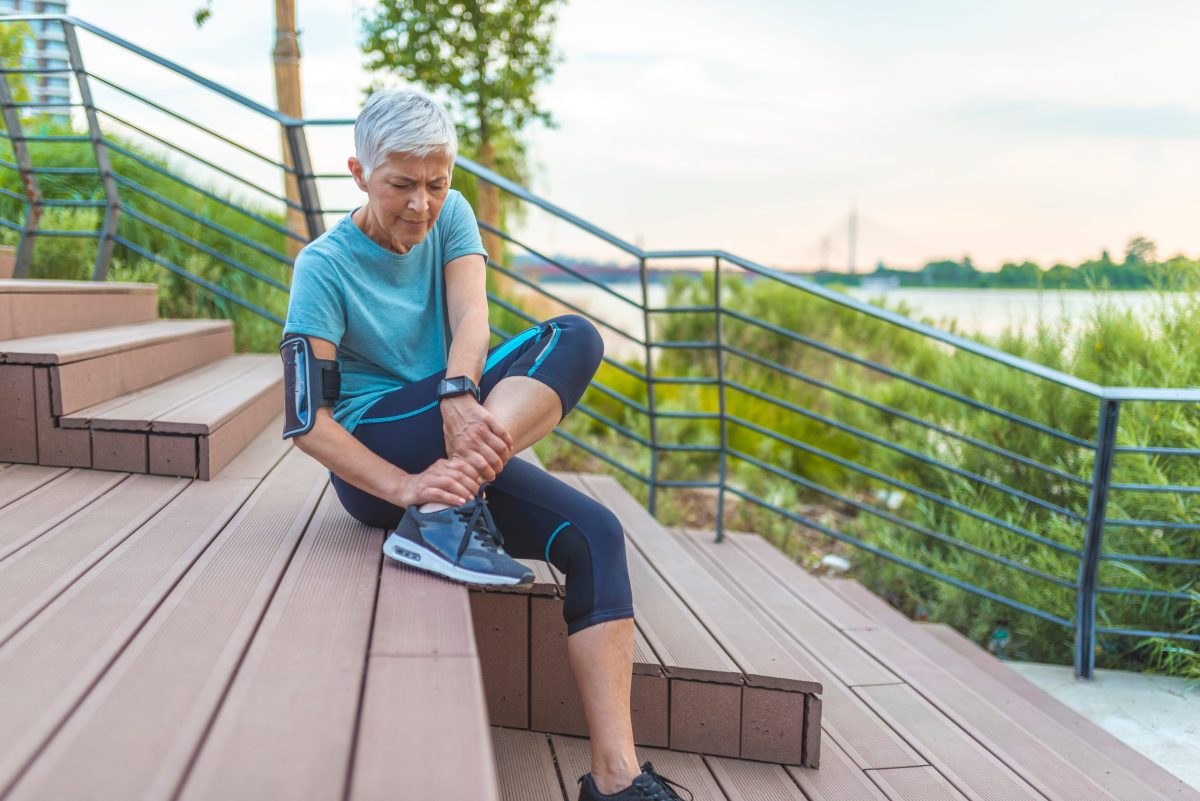A mature woman in workout clothing grapes her ankle in pain sitting on stairs outside by a lake with greenery. 