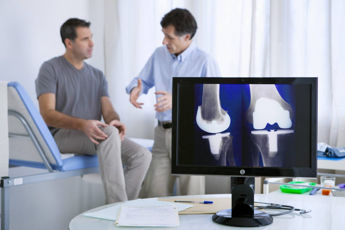 A middle-aged male patient receives a knee exam from an orthopedic surgeon; an image of a knee-replacement is on a computer.