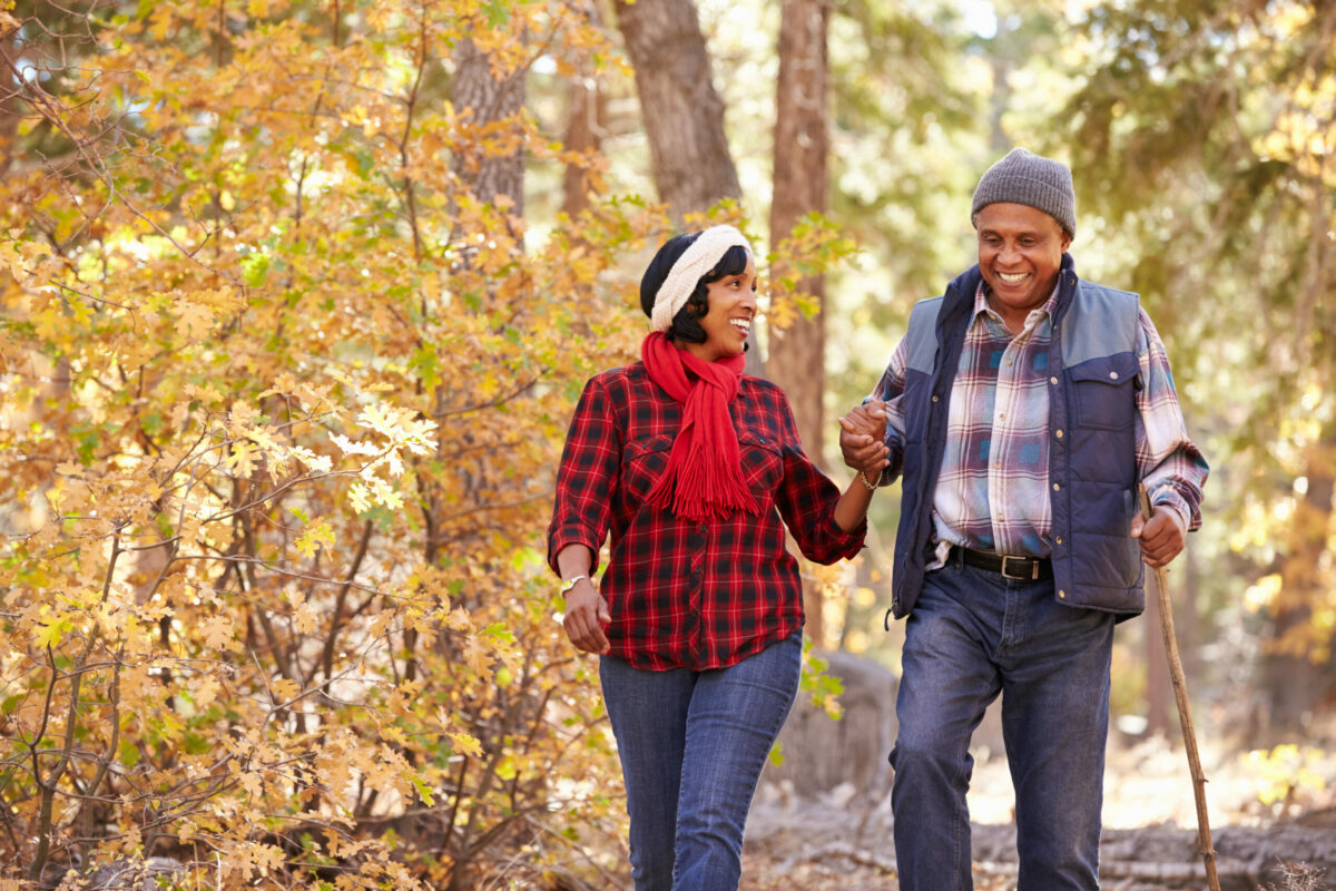 A middle-aged couple walks in the woods holding hands with him using a walking stick after hip preservation. 