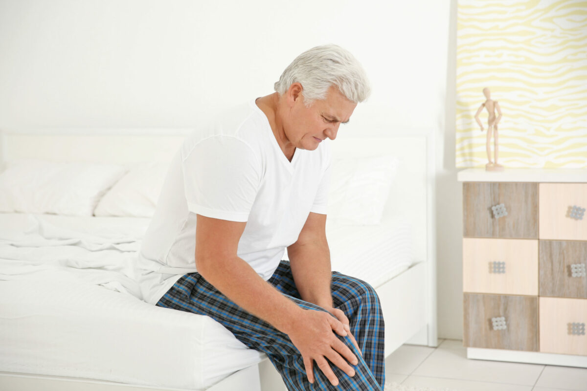  Image of senior man holding a painful knee with both hands while sitting on the edge of the bed. 