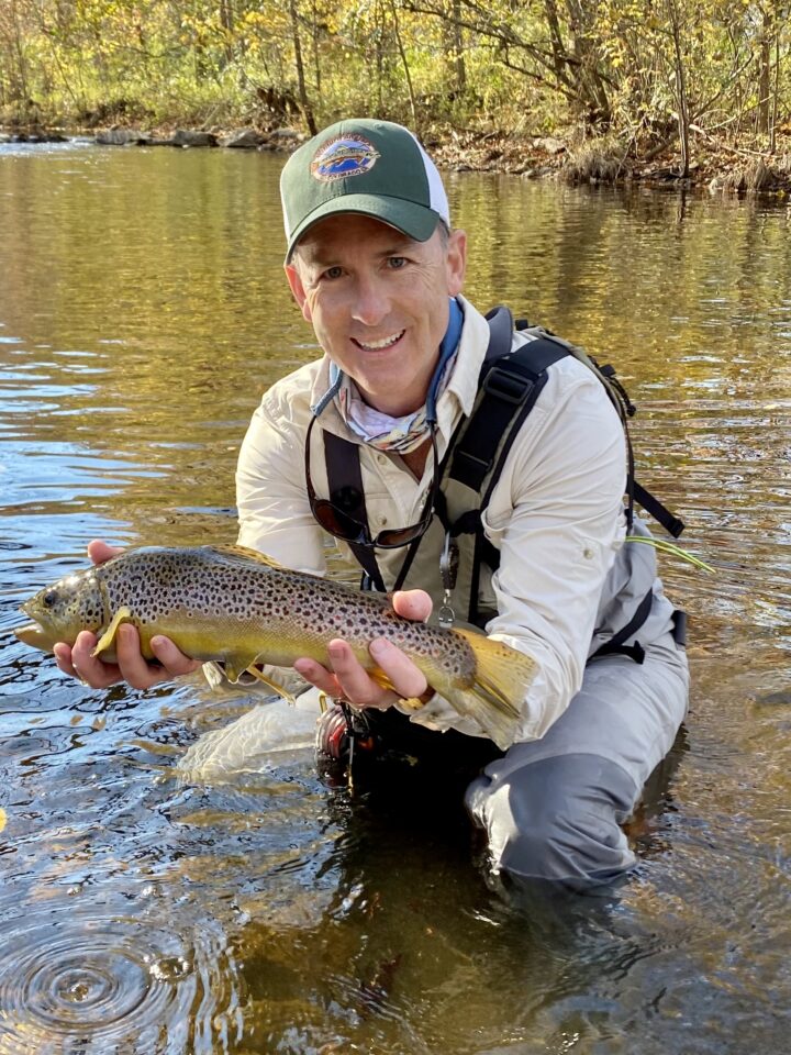 Dr. Mack Aldridge of EmergeOrtho–Triangle wearing a green hat and holding a trout in a river. 