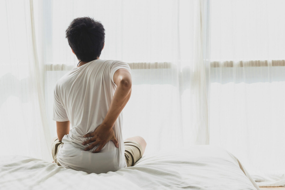 A man holds his lower back in pain while sitting on a bed, needing a back doctor. 