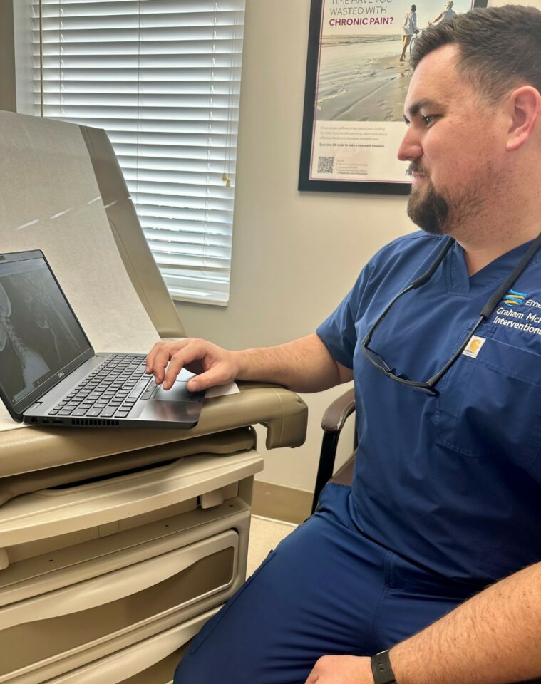 A doctor sits next to an exam table while using a laptop computer.