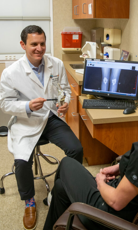 An orthopedic doctor explains a knee replacement using a model and an X-ray on a computer to an attentive patient in a medical office.